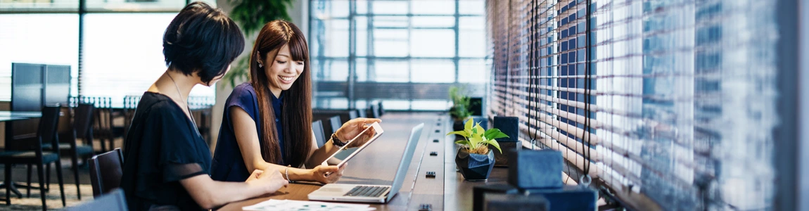 Two young businesspeople seated in a brightly-lit modern workspace surrounded by trees viewing both a laptop and tablet device