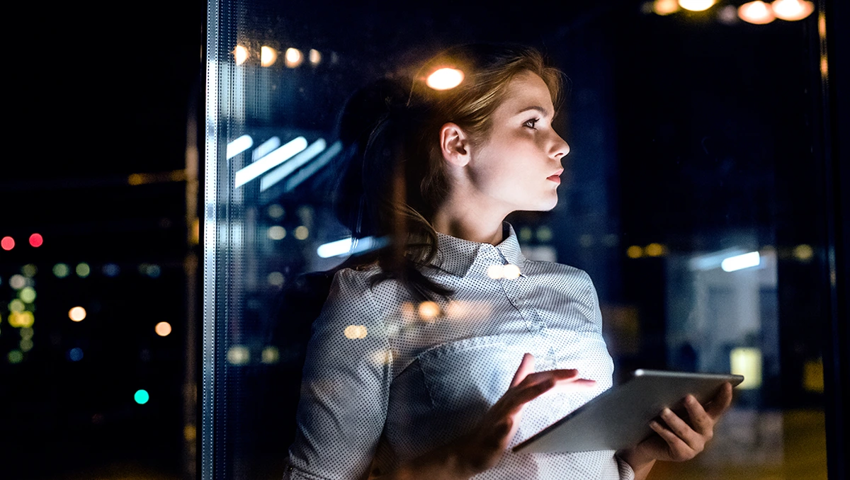 Worried-looking young IT Security professional holding a digital tablet device while staring out of a window in a nighttime modern office space