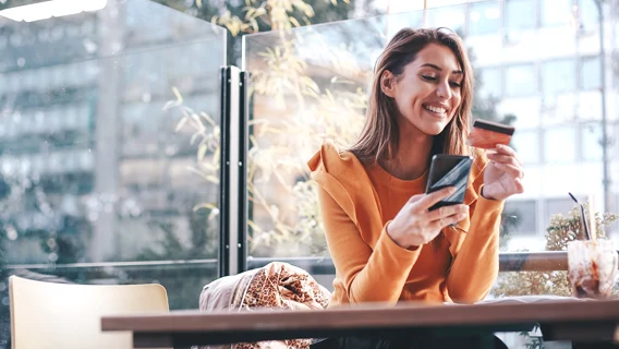 Young woman seated in modern dining environment smiling whilst holding a mobile telephone and payment card