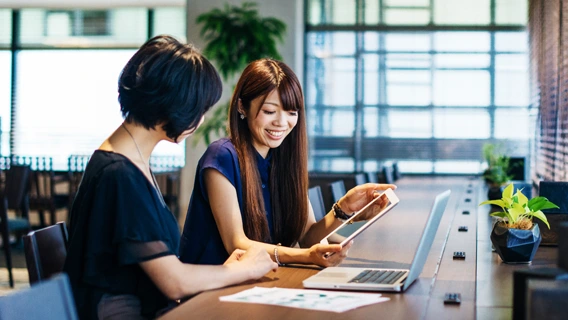 Two young businesspeople seated in a brightly-lit modern workspace surrounded by trees viewing both a laptop and tablet device