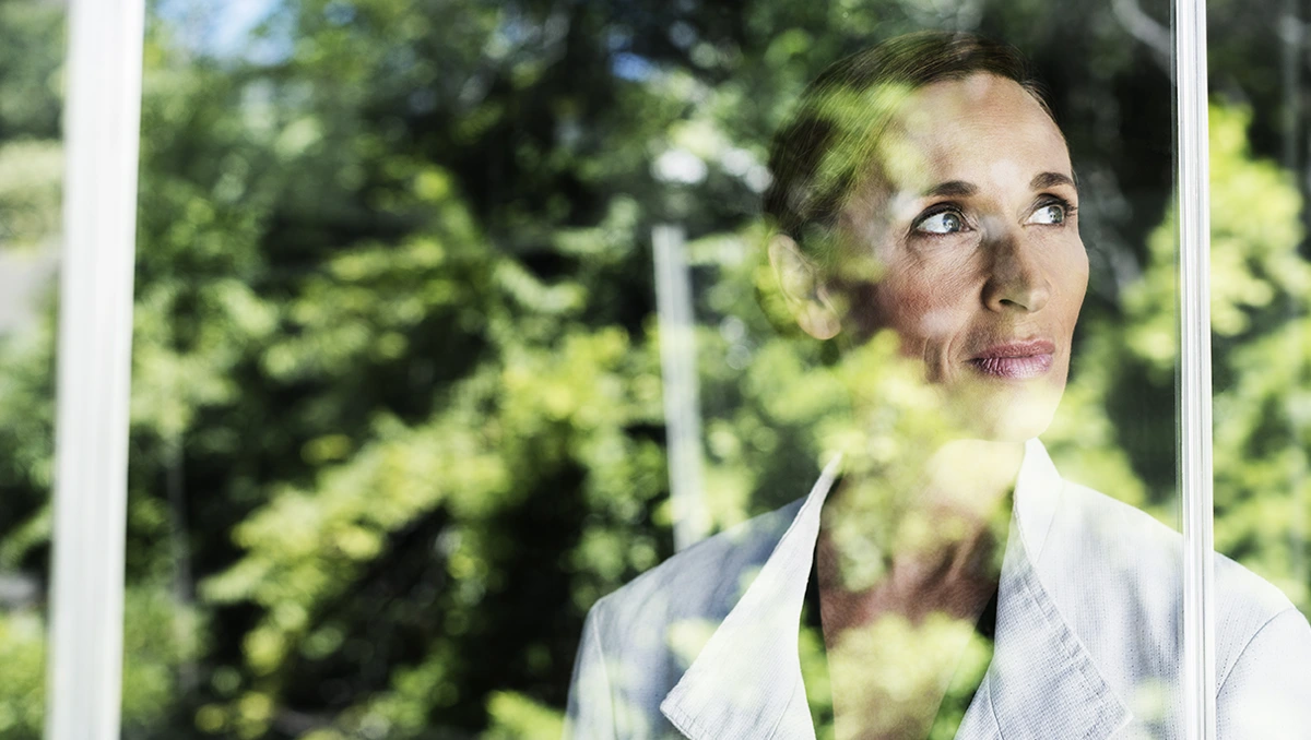 Middle-aged businesswoman staring pensively out of a window that is reflecting lush green leaves on a tree